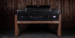 A black Weber gas grill with a lid, set on a custom wooden outdoor table with a dark wood plank wall background.