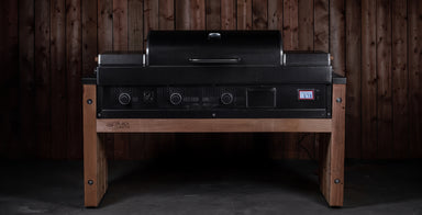 A black Weber gas grill with a lid, set on a custom wooden outdoor table with a dark wood plank wall background.