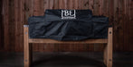 A black grill cover with a white 'BE BLACK EARTH' logo, draped over a wooden grill on a wooden bench against a dark wood plank wall.