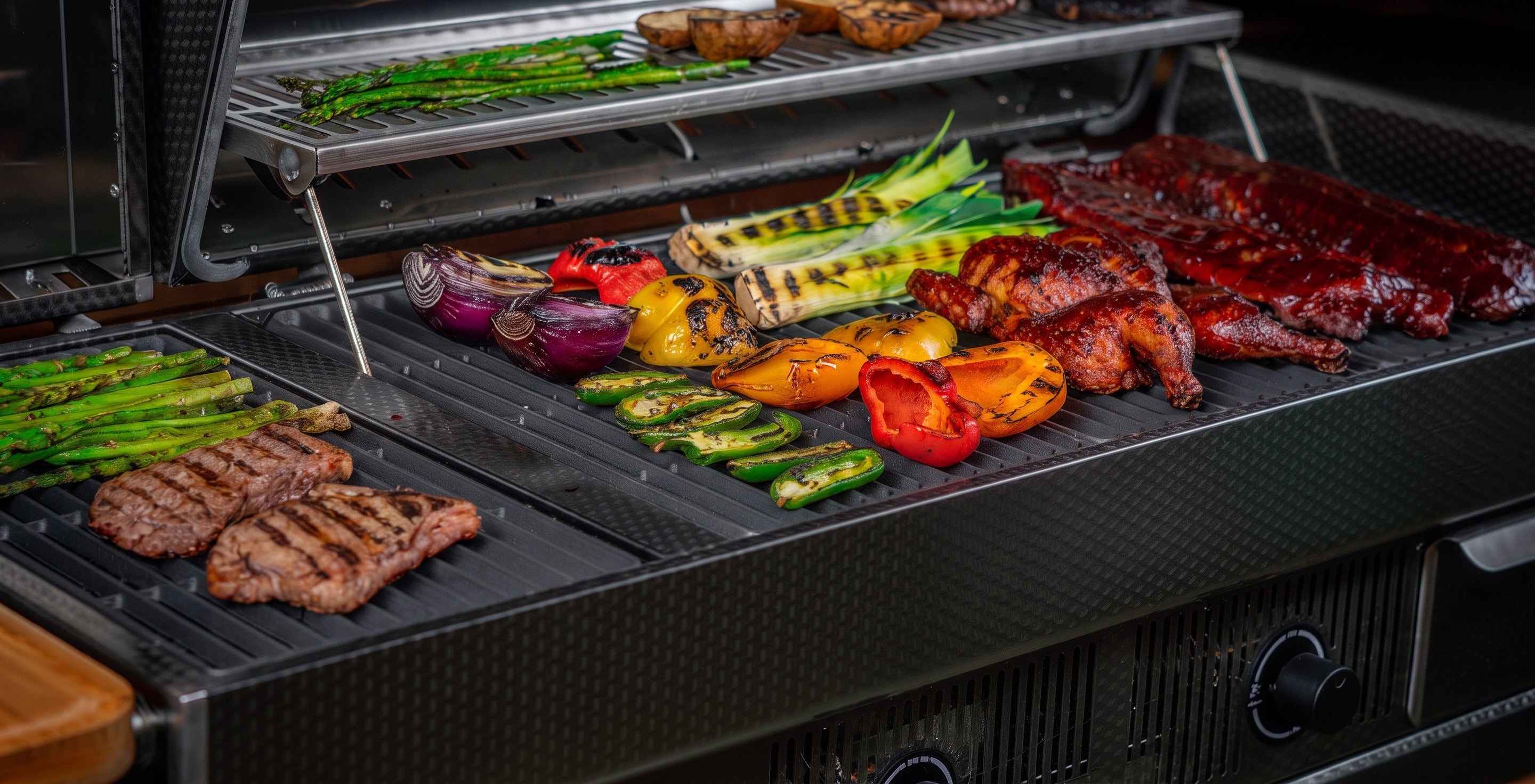 An open charcoal grill with multiple foods cooking on the grates, including ribs, steaks, asparagus, peppers, and corn, shown in a close-up product-style shot.