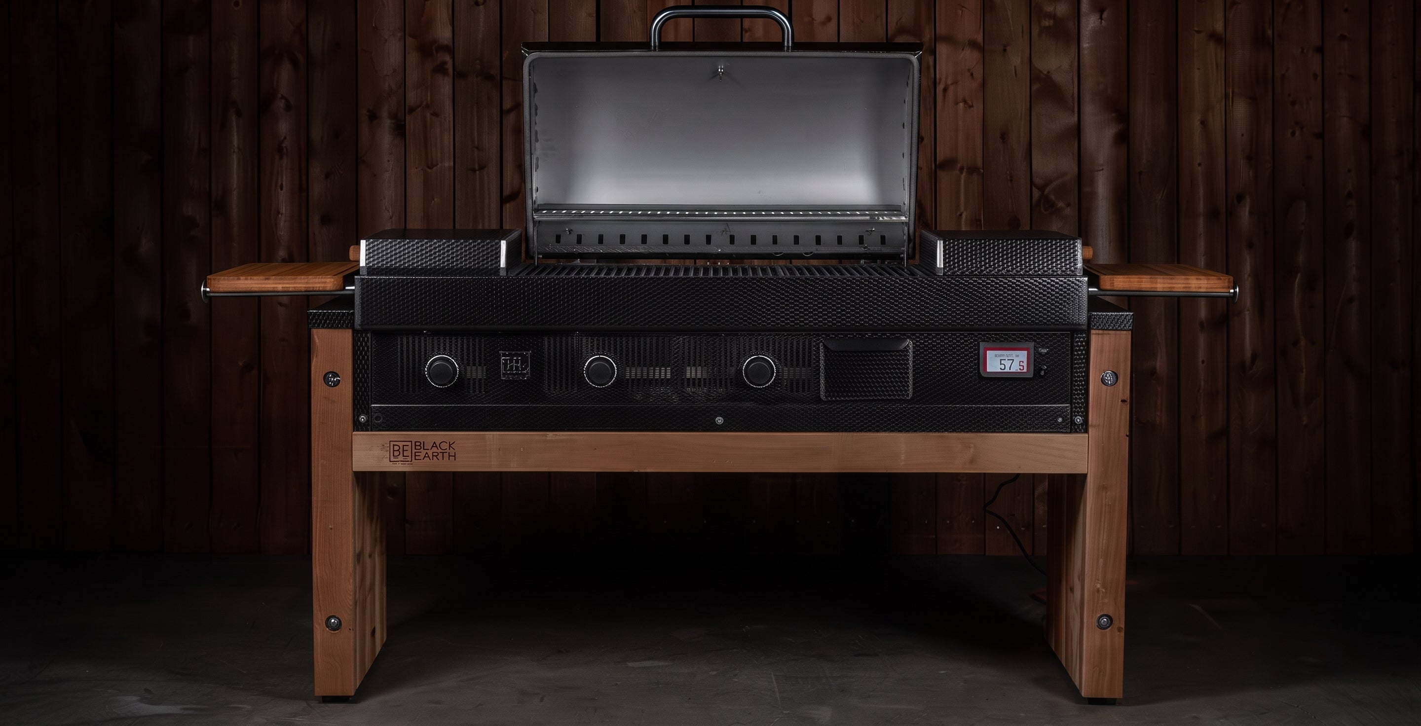 A black outdoor gas grill with the lid open, mounted on a wooden table with side shelves, set against a dark wood plank wall.