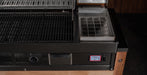 Close-up of a black grill or smoker with a digital temperature display reading 573 on the front panel, alongside a small metal tray and dark interior compartments.