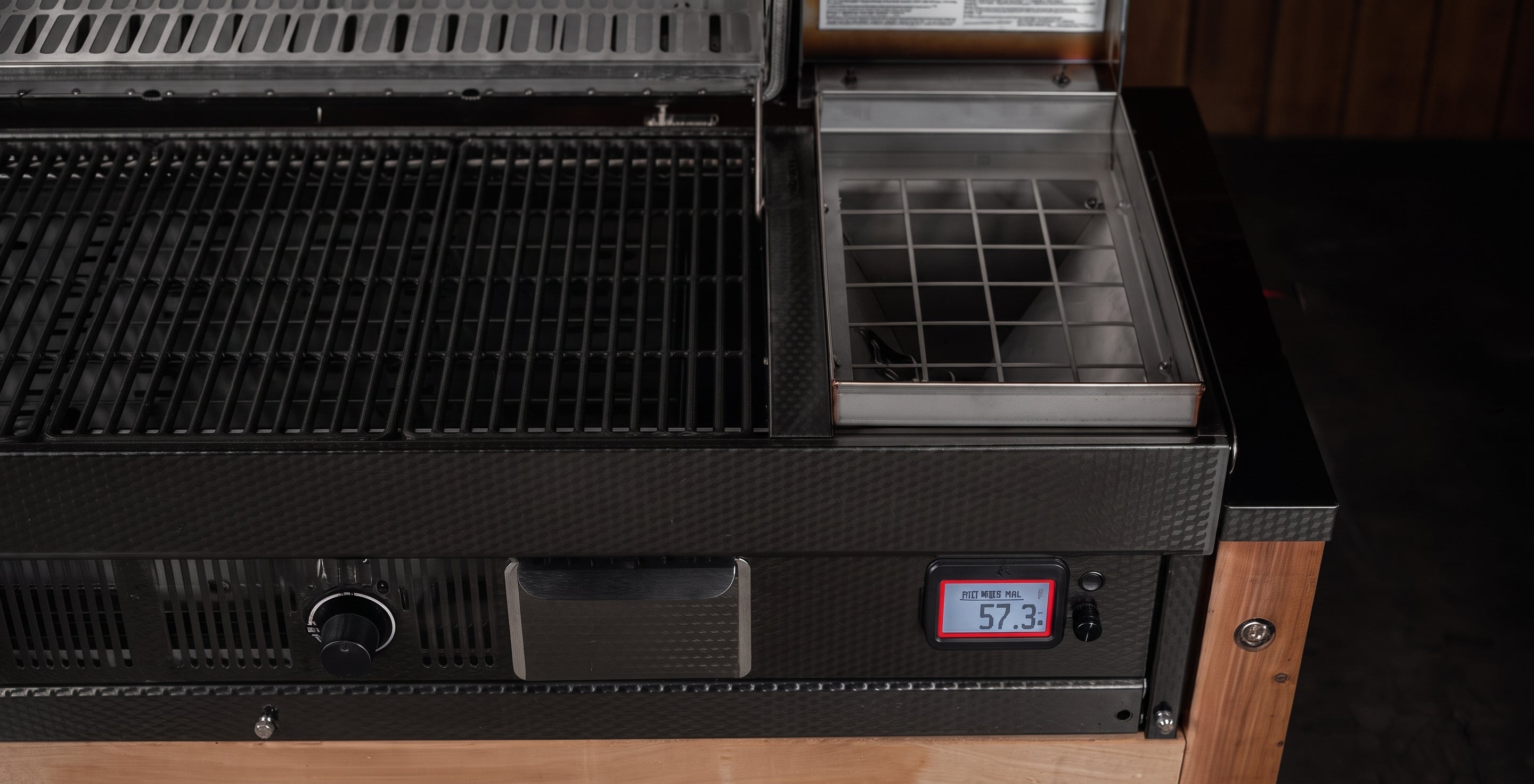 Close-up of a black grill or smoker with a digital temperature display reading 573 on the front panel, alongside a small metal tray and dark interior compartments.