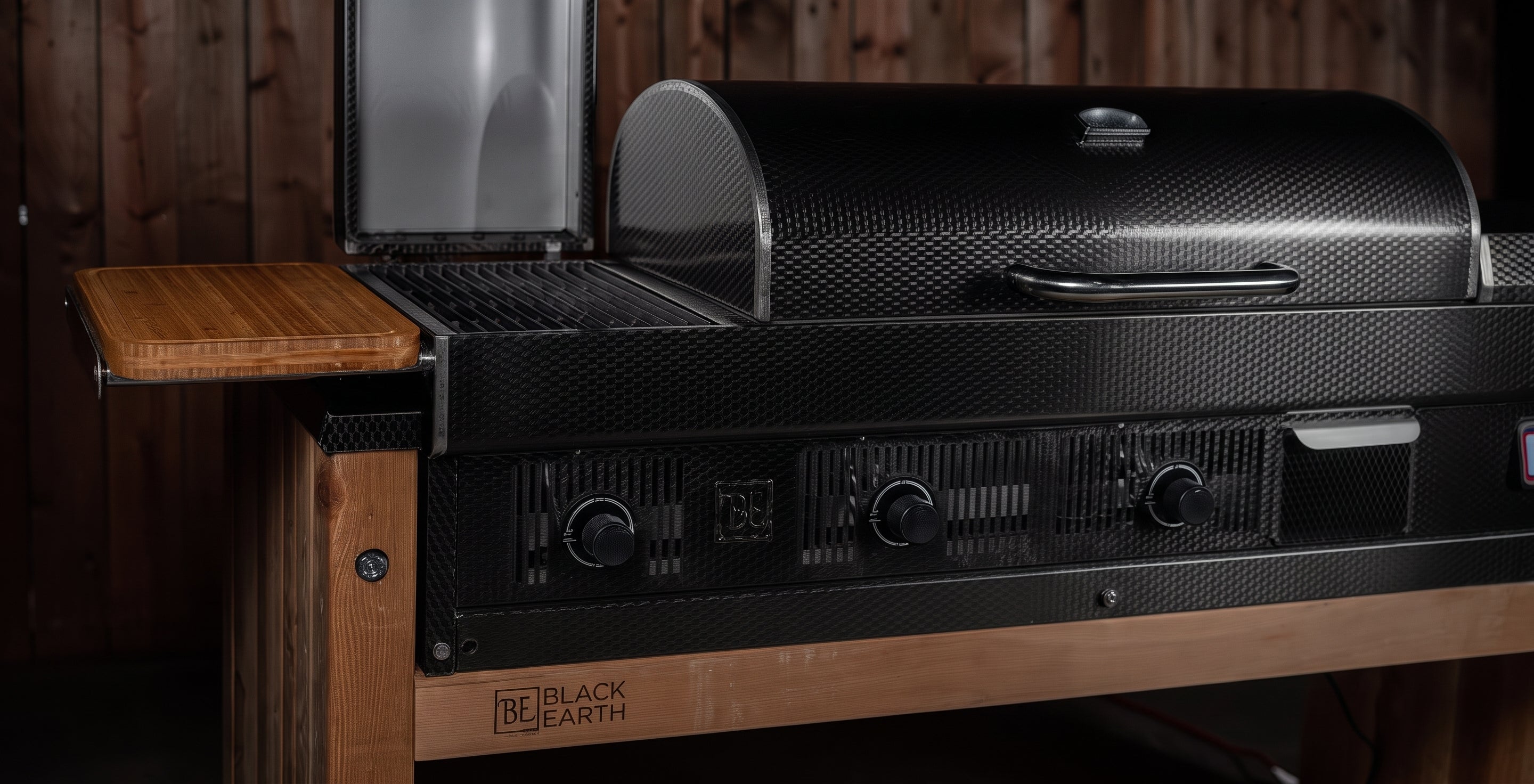 A black carbon-look gas grill with a side table extension, shown in a rustic setting against a dark wood plank wall. The brand name 'Black Earth' is visible on the front wooden base.