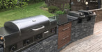 A stainless steel outdoor gas grill with a black lid sits on a stone-tile and wood deck next to matching built-in grills and a black countertop in a backyard setting.