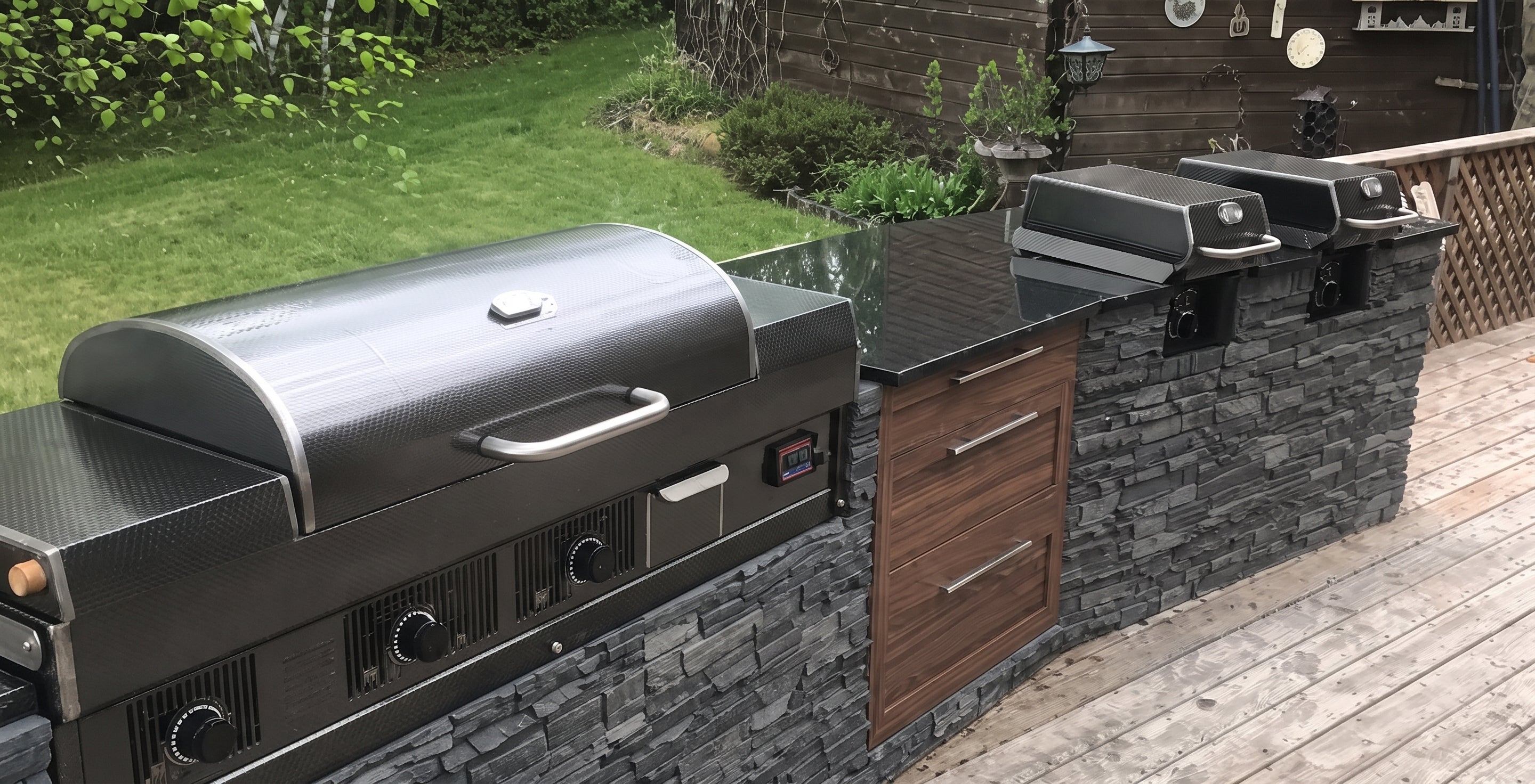 A stainless steel outdoor gas grill with a black lid sits on a stone-tile and wood deck next to matching built-in grills and a black countertop in a backyard setting.