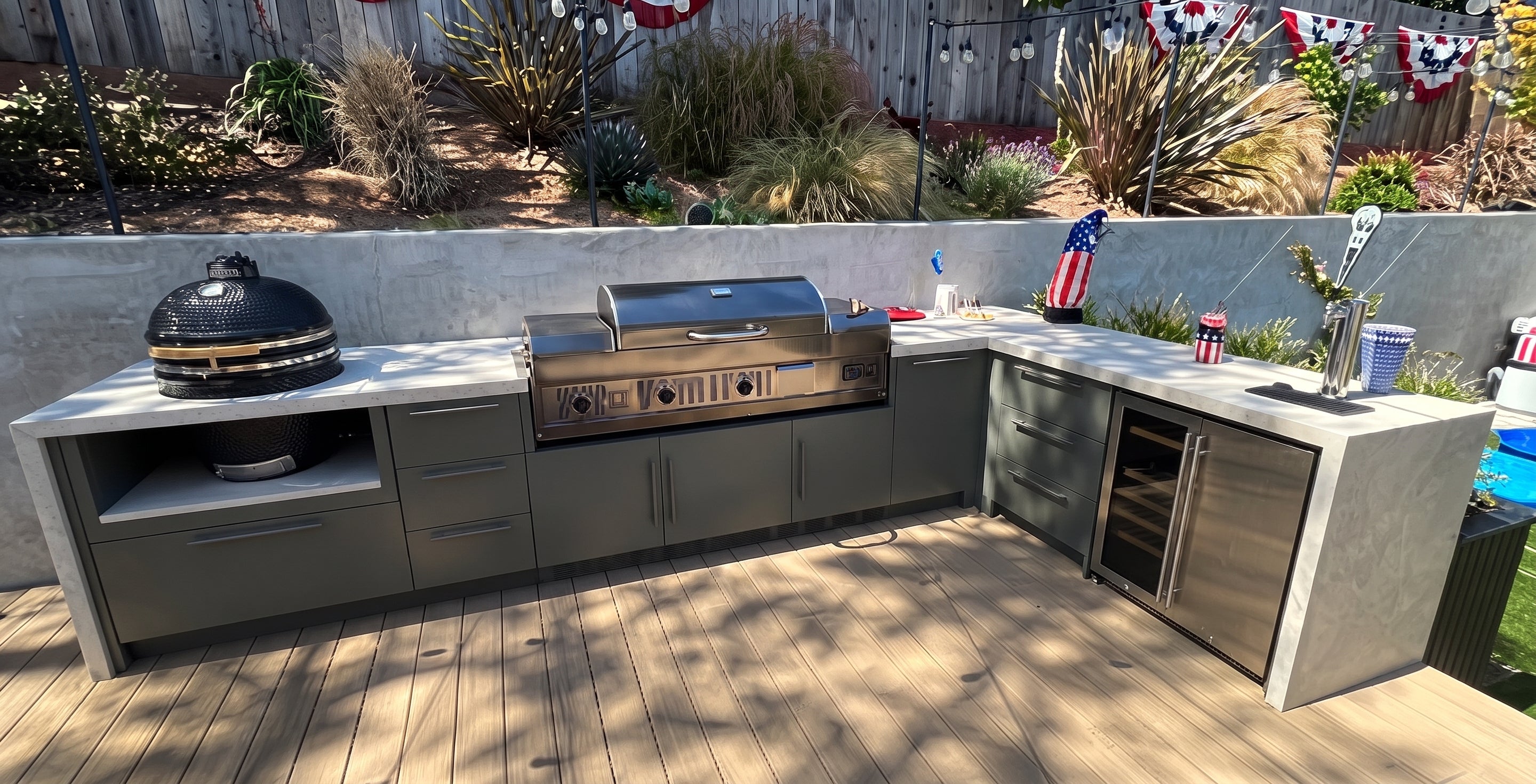 A stainless steel outdoor kitchen island set on a wooden deck, featuring a built-in gas grill, a black dome kamado grill, a small wine cooler, and a concrete countertop with a sink and storage cabinets, set in a backyard with a concrete retaining wall and landscaping behind it.