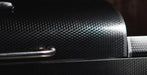 Close-up of a black carbon fiber textured surface with a curved metallic handle and a bright reflective highlight.