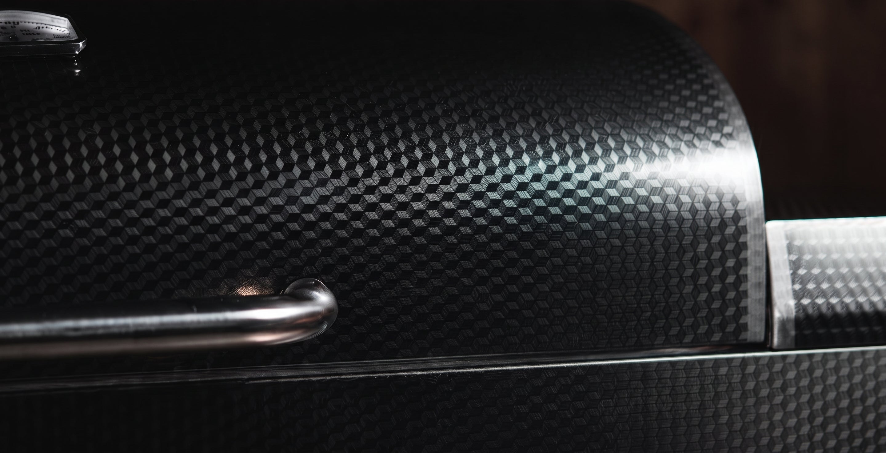 Close-up of a black carbon fiber textured surface with a curved metallic handle and a bright reflective highlight.