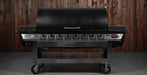 A black and stainless steel outdoor gas grill with six burners and a lid, shown against a rustic wooden plank wall background.
