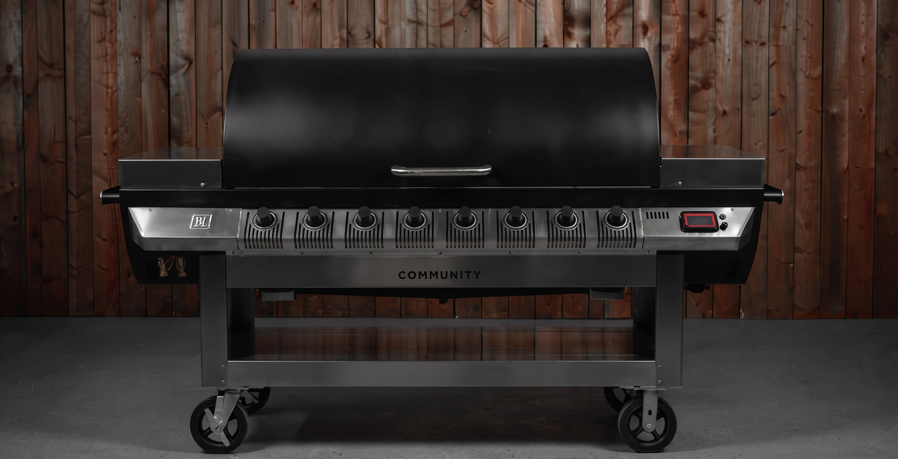 A black and stainless steel outdoor gas grill with six burners and a lid, shown against a rustic wooden plank wall background.