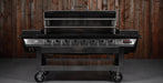 A large black and stainless steel gas grill with six burners and a lid handle, shown with the lid open against a dark wood plank wall.