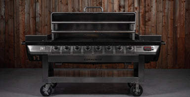 A large black and stainless steel gas grill with six burners and a lid handle, shown with the lid open against a dark wood plank wall.