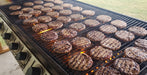 Multiple grilled hamburger patties cook on a large outdoor gas grill with visible flames and control knobs, with grass in the background.