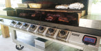 A stainless steel multi-zone smoker grill with six control knobs and a digital temperature display, shown cooking ribs and other meats on a covered patio.