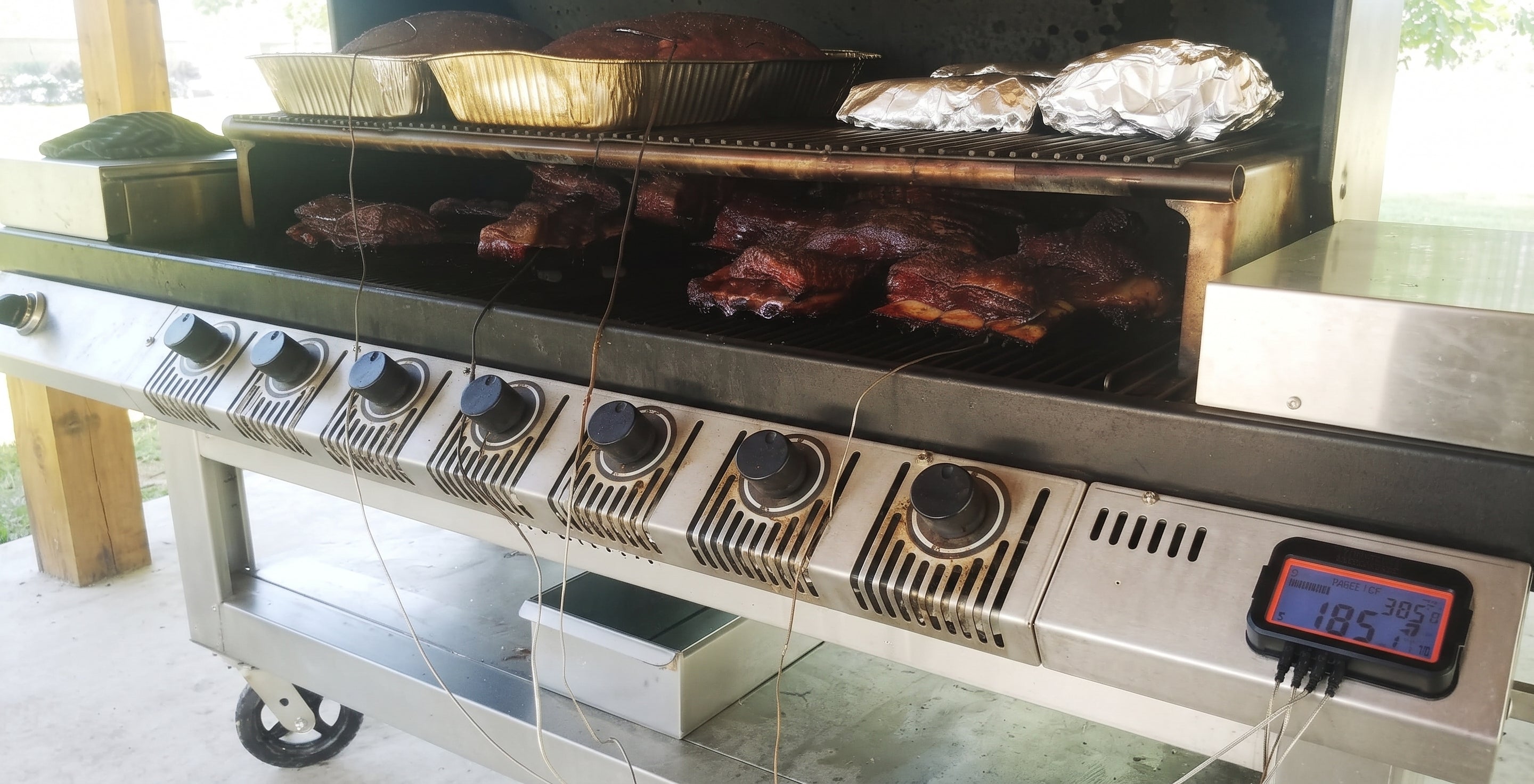 A stainless steel multi-zone smoker grill with six control knobs and a digital temperature display, shown cooking ribs and other meats on a covered patio.