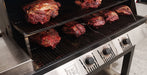 Multiple cuts of raw beef or lamb roasting on the shelves of a stainless steel gas grill with black grates and control knobs.