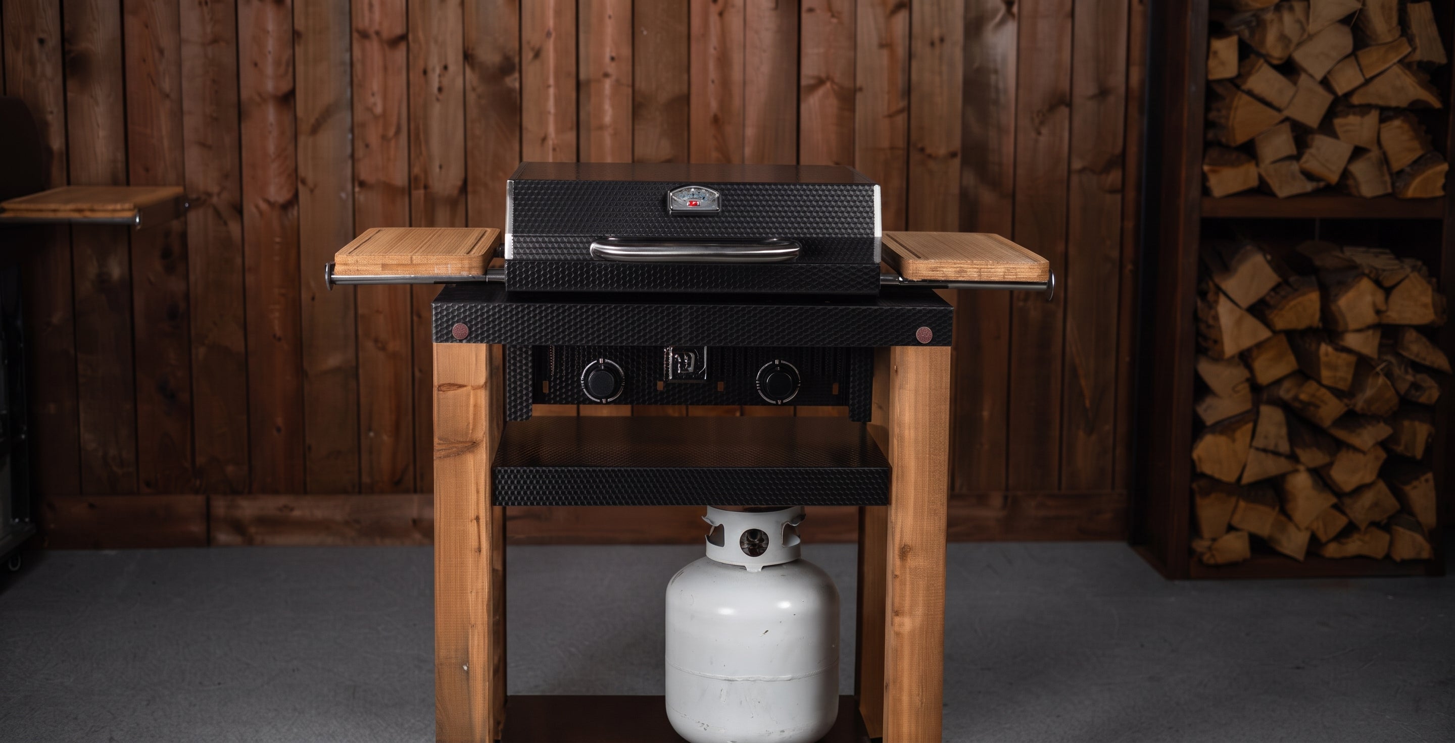 A black gas grill mounted on a rustic wooden stand with side shelves, set against a wood-paneled wall with stacked firewood visible on the right.