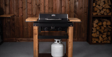 A black gas grill mounted on a rustic wooden stand with side shelves, set against a wood-paneled wall with stacked firewood visible on the right.