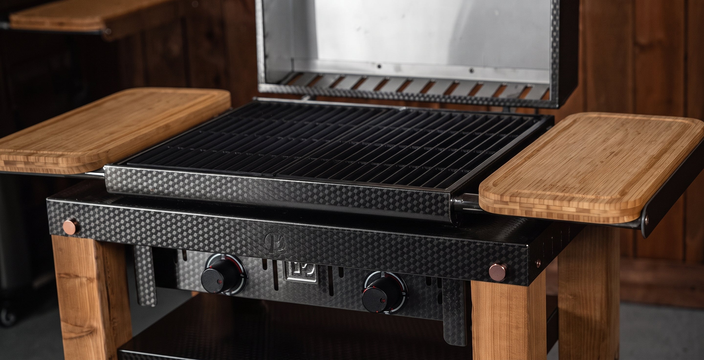 A close-up of a black gas grill with a textured carbon-fiber-look front panel, black grates, and light brown wooden side tables and legs, shown in a rustic indoor setting.