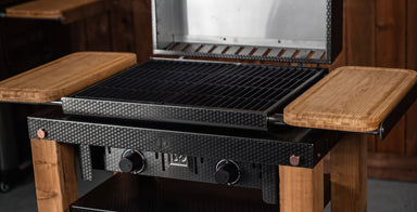 A close-up of a black gas grill with a textured carbon-fiber-look front panel, black grates, and light brown wooden side tables and legs, shown in a rustic indoor setting.