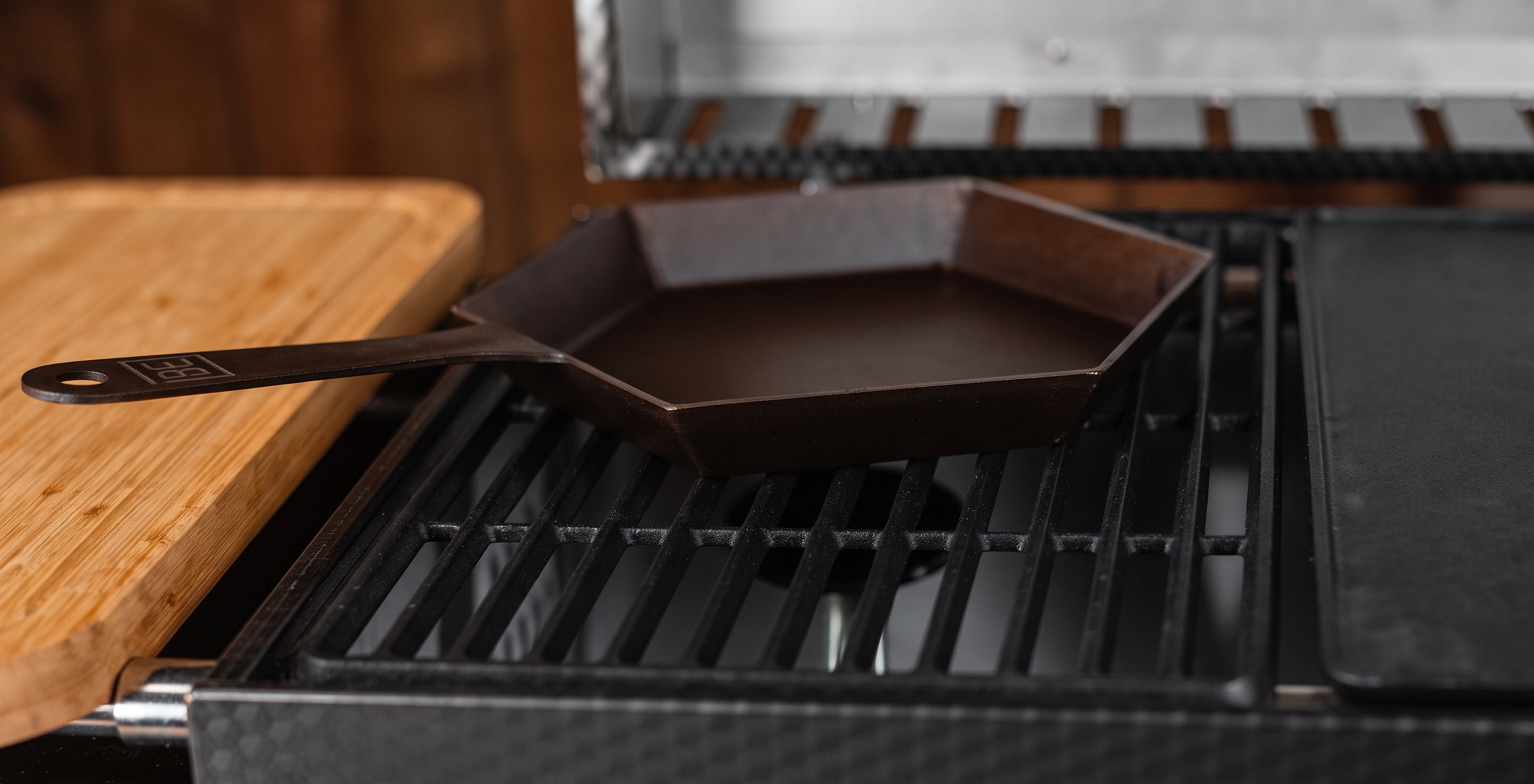 A dark cast iron skillet with a hexagonal shape and a long handle, resting on the grates of a black grill next to a wooden cutting board.