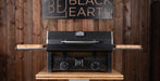 A black carbon-look gas grill with wooden side tables and a metal control panel, displayed on a wooden stand against a wood-paneled wall with a 'Black Earth' sign.