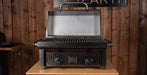 A black gas grill with a textured exterior and a stainless steel interior lid, shown open on a wooden table against a rustic wood plank wall.