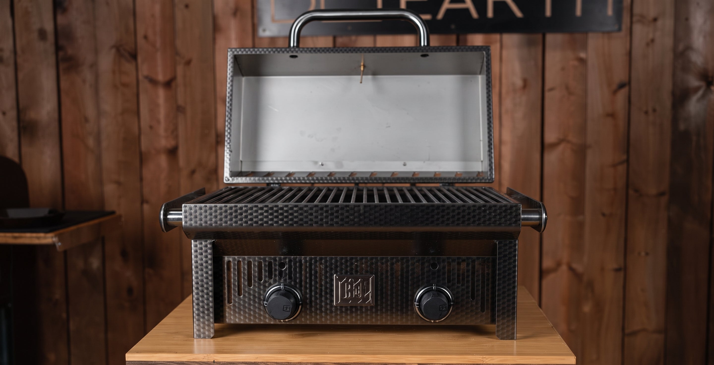 A black gas grill with a textured exterior and a stainless steel interior lid, shown open on a wooden table against a rustic wood plank wall.