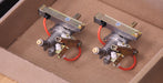 Two small mechanical components with red wires and brass-toned metal parts resting inside a brown cardboard box.