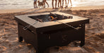 A square black outdoor fire pit table on a sandy beach, with logs burning inside and people and dogs standing in the background near the water at sunset.