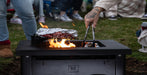 A black outdoor tabletop grill with visible flames and foil-wrapped food, shown in a real outdoor setting with people standing around it. The grill has a visible BECKYARD logo on the front.