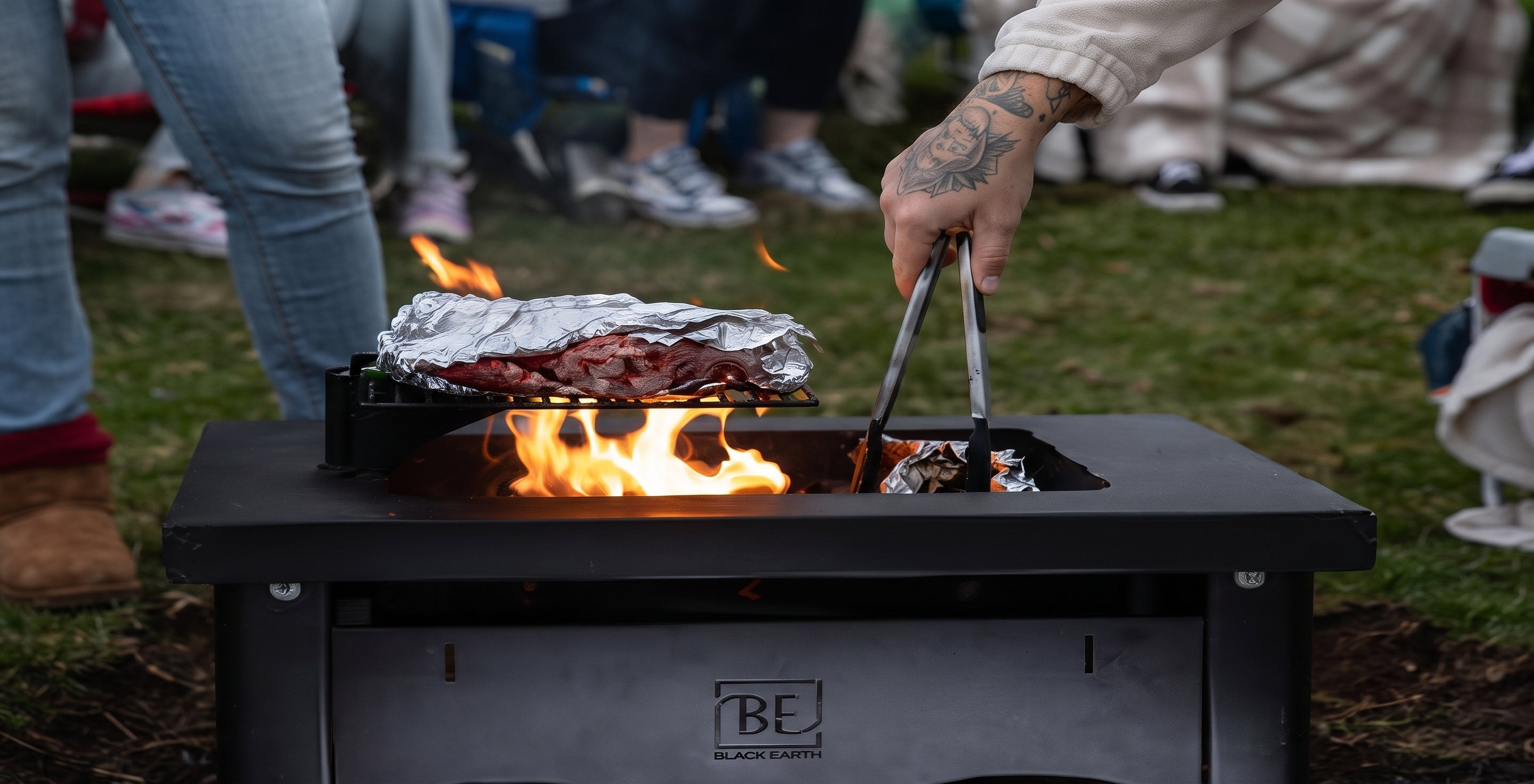 A black outdoor tabletop grill with visible flames and foil-wrapped food, shown in a real outdoor setting with people standing around it. The grill has a visible BECKYARD logo on the front.
