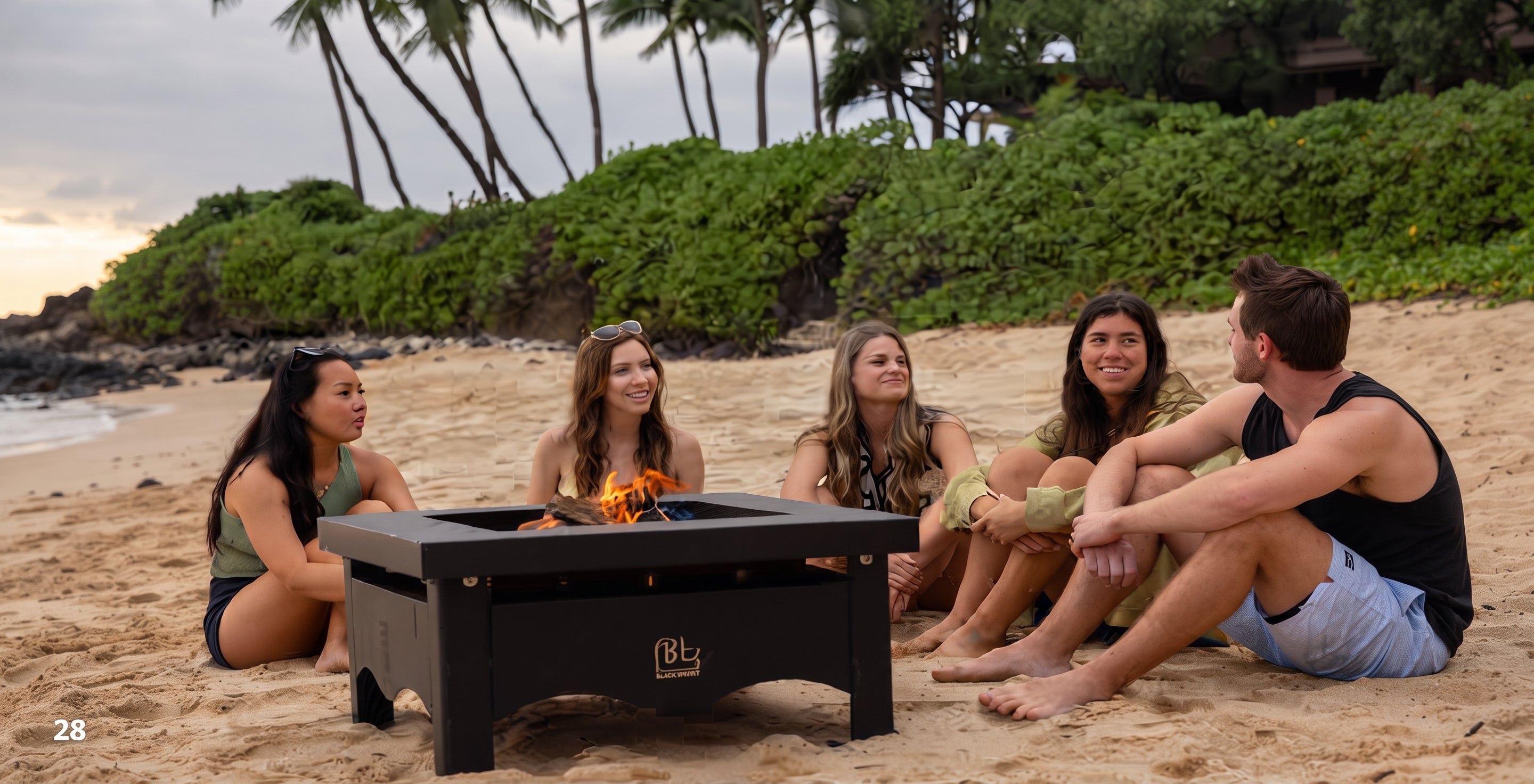 Five people sitting on a sandy beach around a black outdoor fire pit table with a fire burning inside. Palm trees and green foliage are visible in the background under a sunset sky.
