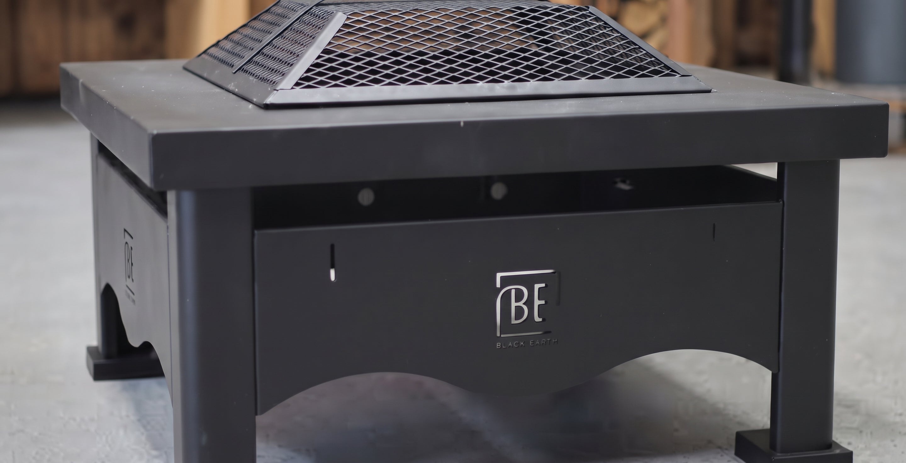 A black outdoor fire pit table with a metal grate top and a visible 'BF Firestone' logo on the front panel, set against a blurred concrete and wood background.