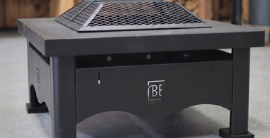 A black outdoor fire pit table with a metal grate top and a visible 'BF Firestone' logo on the front panel, set against a blurred concrete and wood background.