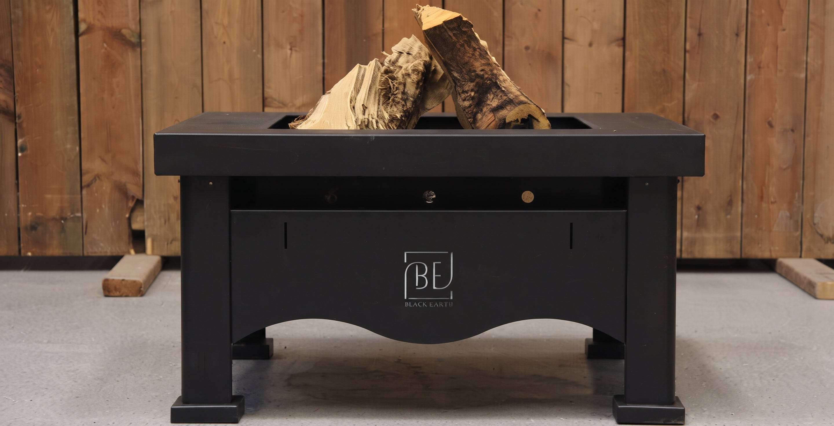 A black outdoor fire pit table with a built-in wood fire and stacked logs, featuring a front drawer and a 'BE BLACK ESTATE' logo, set against a rustic wooden plank wall.