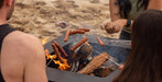 A black metal fire pit table on a sandy beach, with people roasting sausages over an open fire using metal skewers.