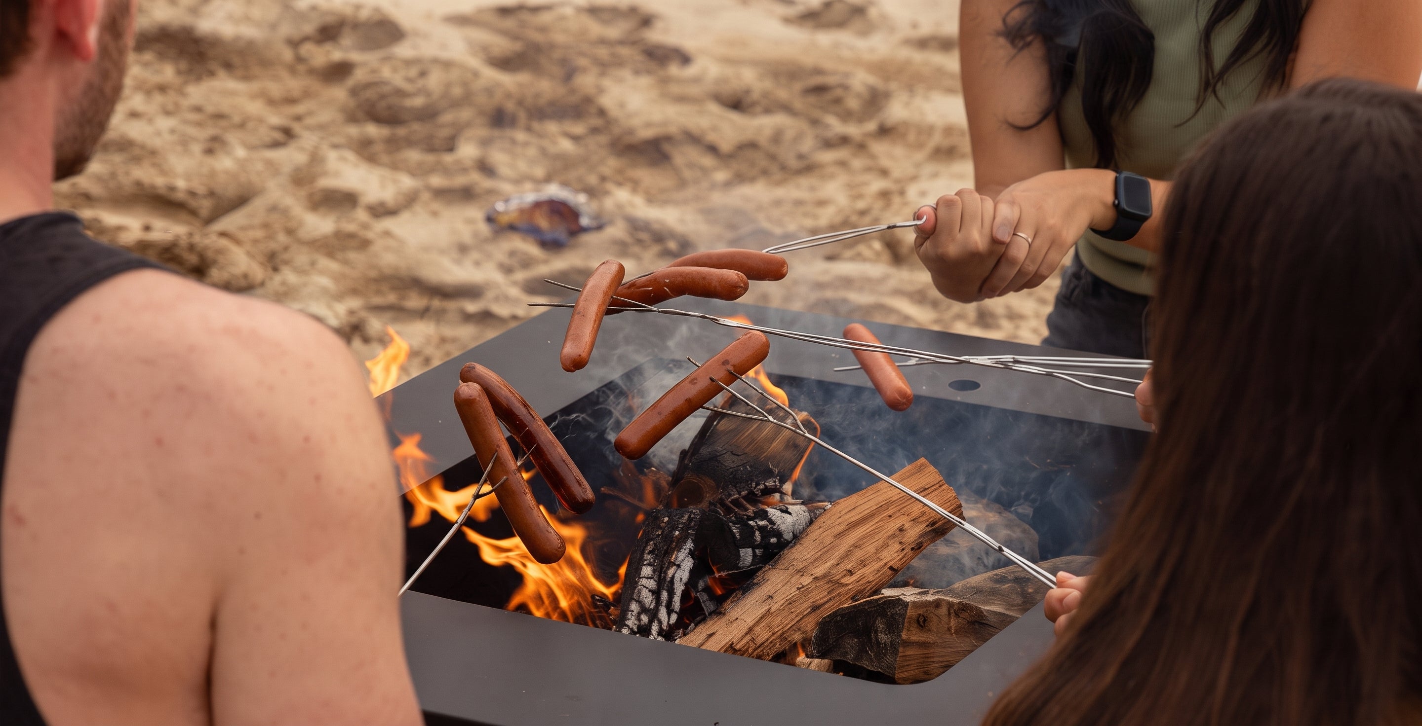 A black metal fire pit table on a sandy beach, with people roasting sausages over an open fire using metal skewers.