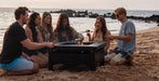 A group of five friends gathered around a black outdoor fire pit table on a sandy beach at sunset, with the ocean and dark rocks in the background.