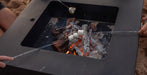 A square black metal fire pit table on a sandy beach, with marshmallows roasting over embers and visible human hands holding the roasting sticks.