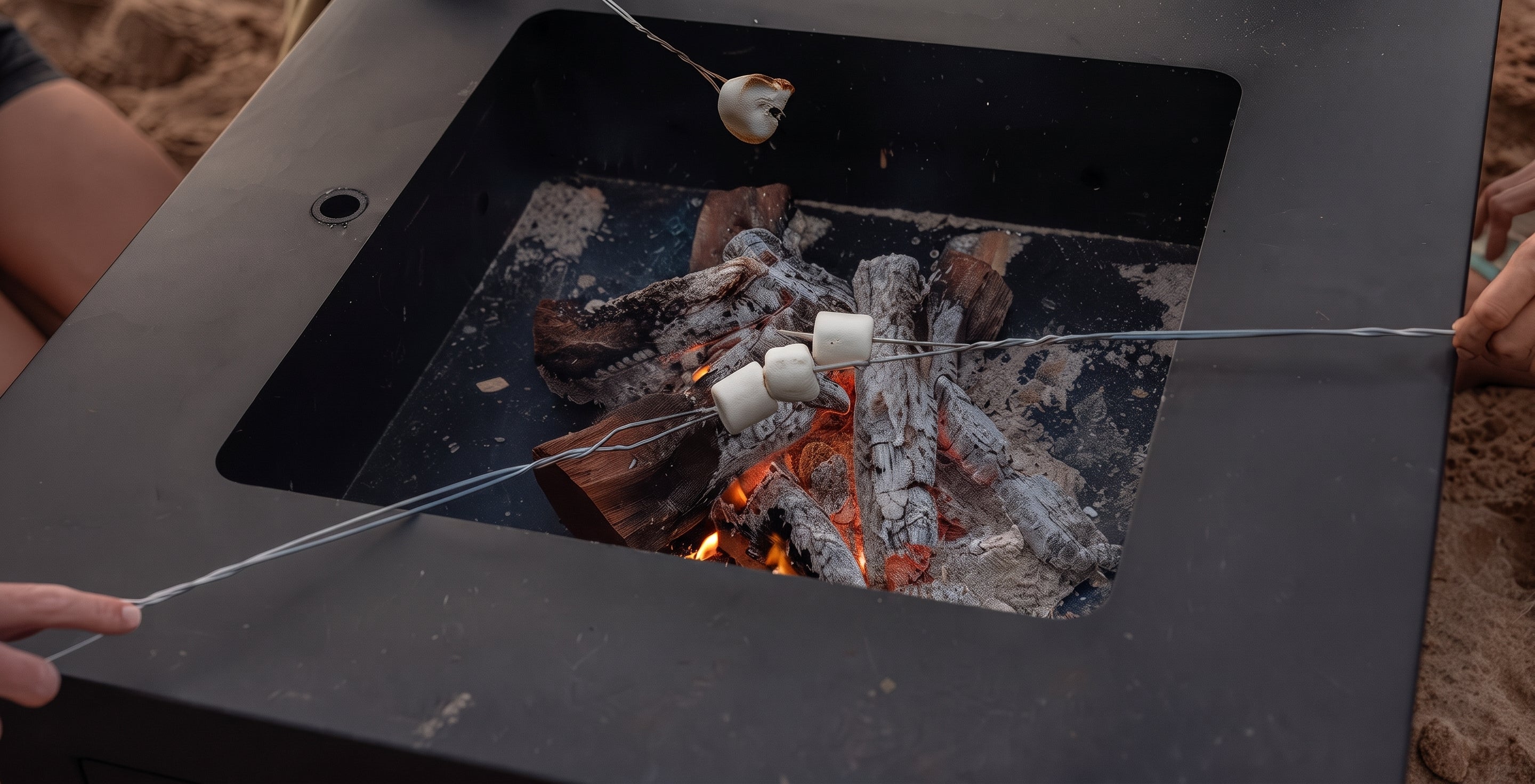 A square black metal fire pit table on a sandy beach, with marshmallows roasting over embers and visible human hands holding the roasting sticks.