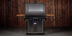 A stainless steel gas grill with side tables and a lid handle, shown open against a dark wood plank wall background.