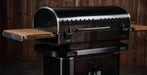 A black gas grill with a curved lid, stainless steel handle, and two wooden side tables extended on either side, set against a dark wood plank background.