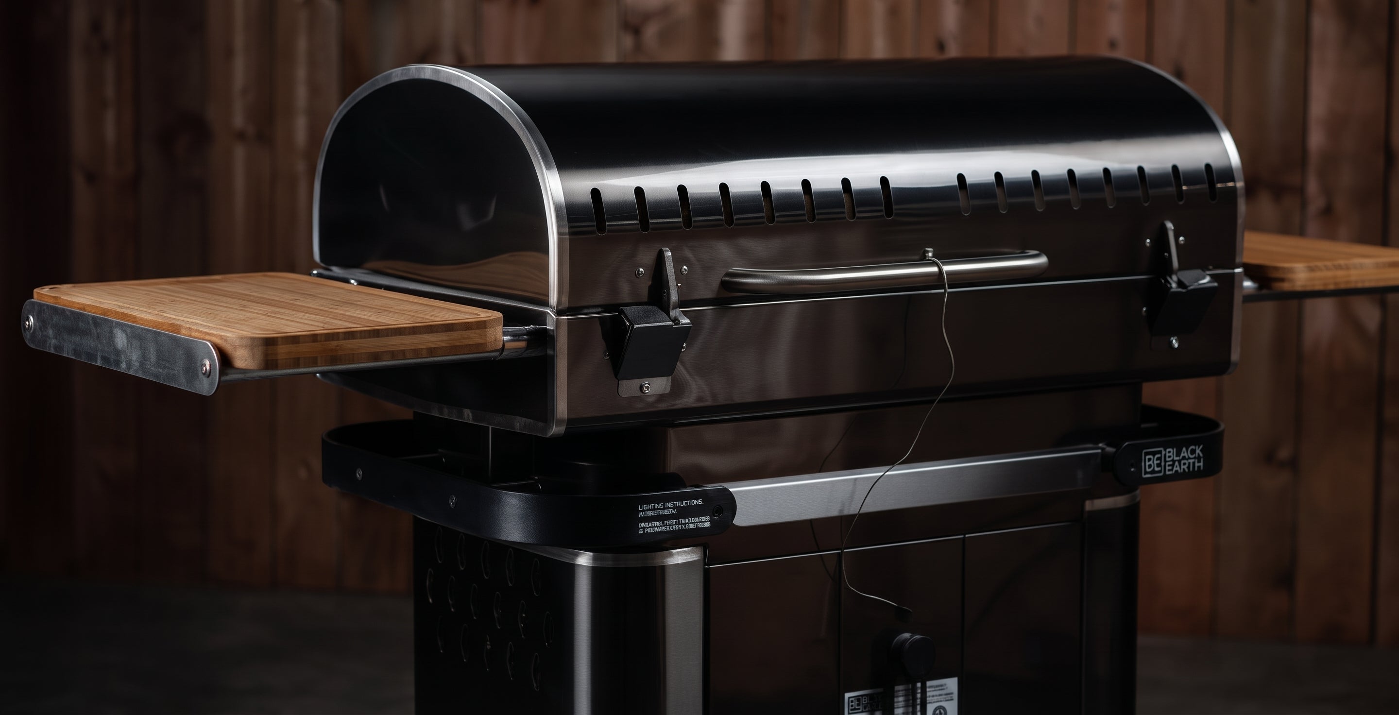 A black gas grill with a curved lid, stainless steel handle, and two wooden side tables extended on either side, set against a dark wood plank background.