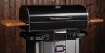 A black and stainless steel outdoor grill with wooden side shelves, set against a dark wood plank wall background.
