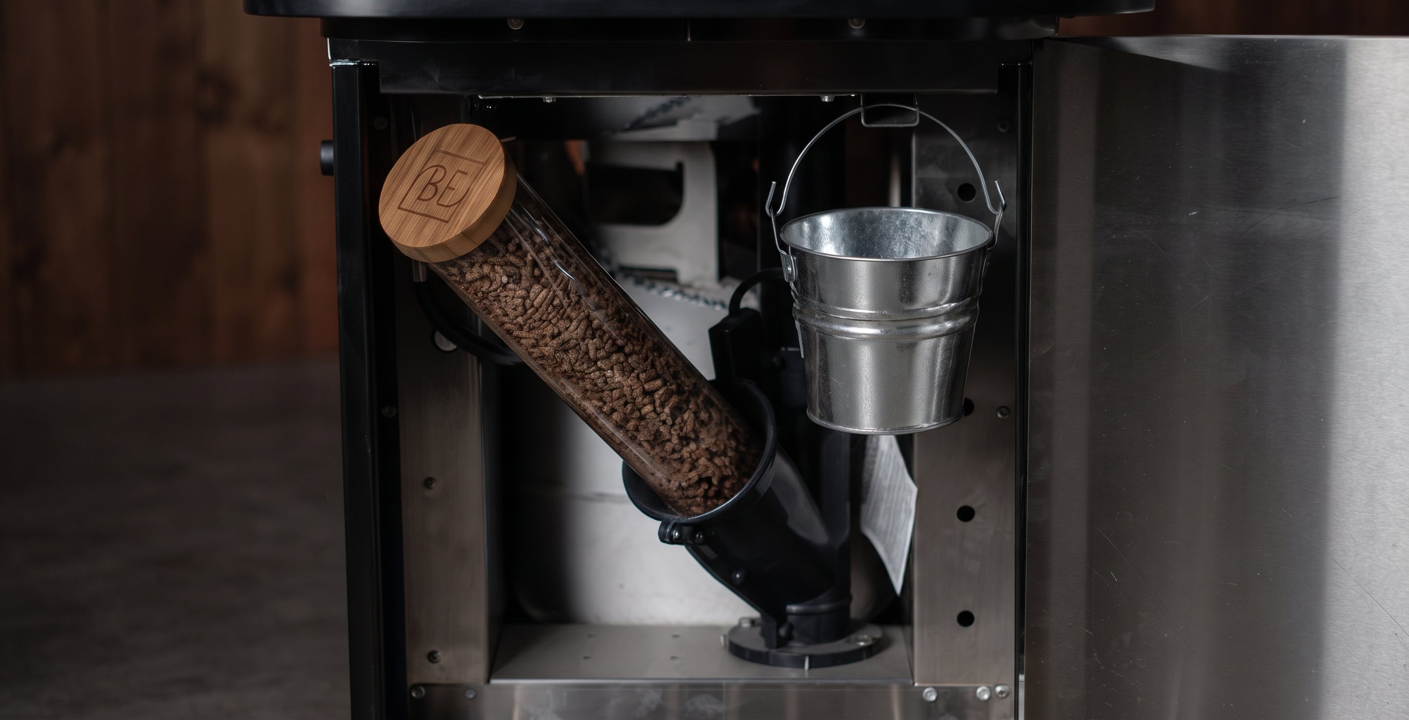 Close-up of the interior of a metal outdoor fireplace or stove, showing a cylindrical wood-burning chamber with a wooden lid, a metal bucket hanging on the side, and a burner assembly inside.