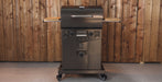 A charcoal grill with side tables and a wheeled base, shown against a wooden plank wall.