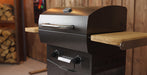 A black outdoor gas grill with a textured finish, featuring a silver handle, control knobs, and two light wood side tables, set against a rustic wooden wall and blurred background.