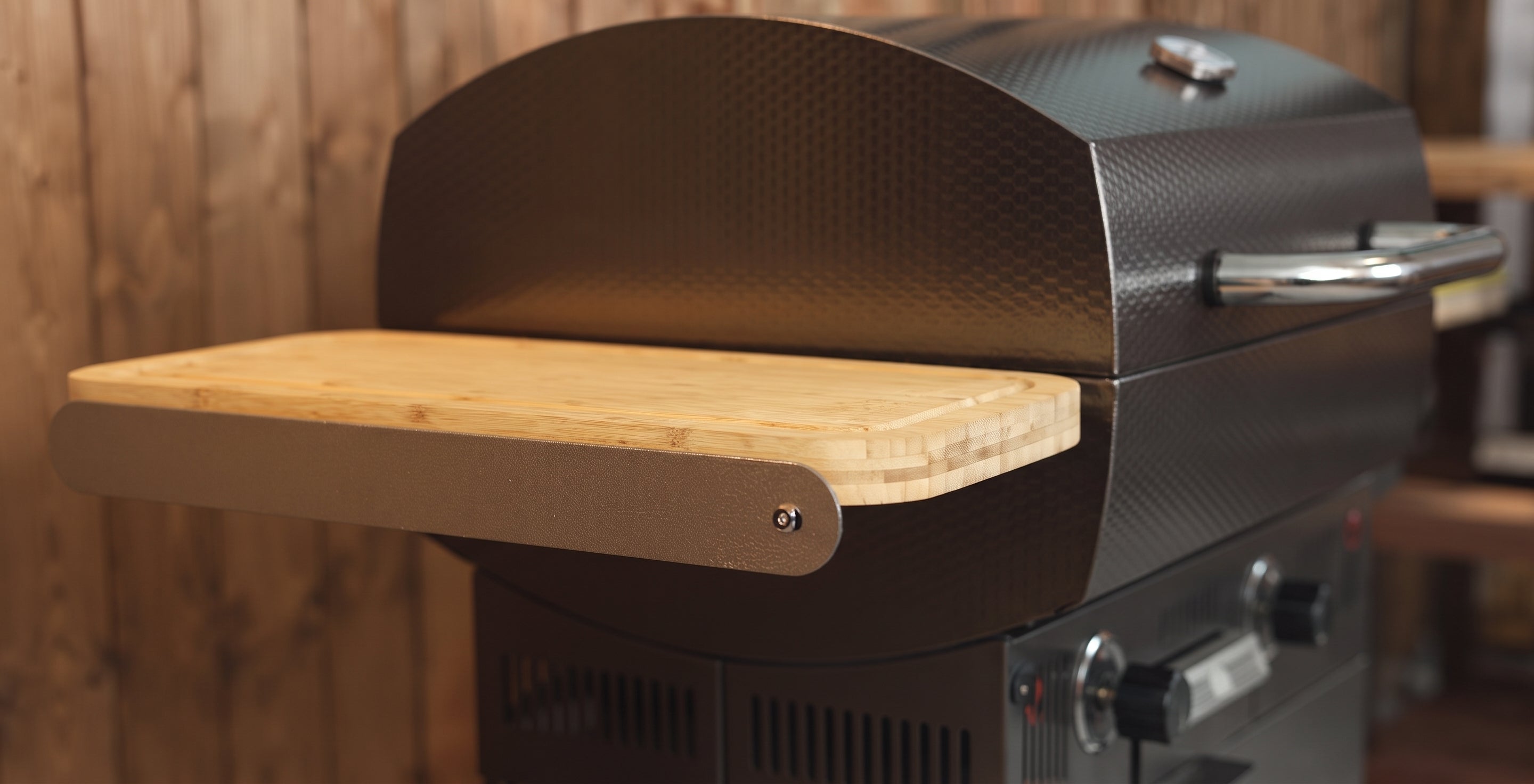 A close-up of a black gas grill with a wooden side table extension, set against a rustic wood plank background.